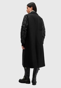 Lungo cappotto nero con maniche a sbuffo nere a contrasto, caratterizzato da una texture liscia e una silhouette strutturata. Abbinato a stivali neri fino al ginocchio.