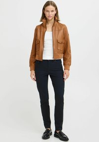 Giovane donna in piedi che indossa una giacca in pelle beige, una canotta bianca, pantaloni neri aderenti e mocassini neri, davanti a uno sfondo chiaro e semplice.
