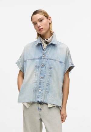 Vrouw draagt een lichtgewassen, oversized spijkerjack met korte mouwen, rafelige randen en lichte spijkerbroek, staand tegen een effen achtergrond.