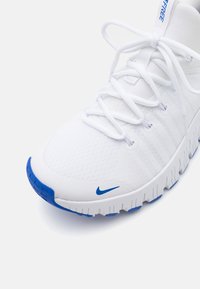 Vit sportskor med texturerad mesh-ovandel, blå Nike-logotyp och en flexibel sula med ett vågigt mönster. Snörningen är vit.