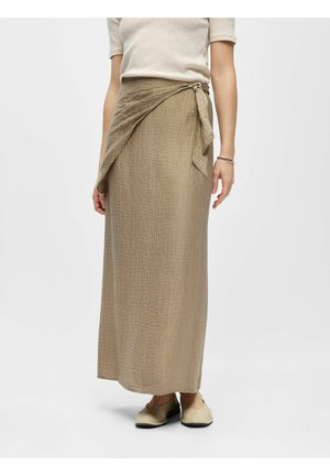 Femme portant une jupe portefeuille texturée beige, un haut crème à manches courtes et des chaussures plates beige clair sur un fond uni.