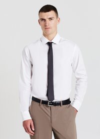 Homme portant une chemise blanche, une cravate noire et un pantalon beige avec une ceinture noire, se tenant devant un fond clair uni.