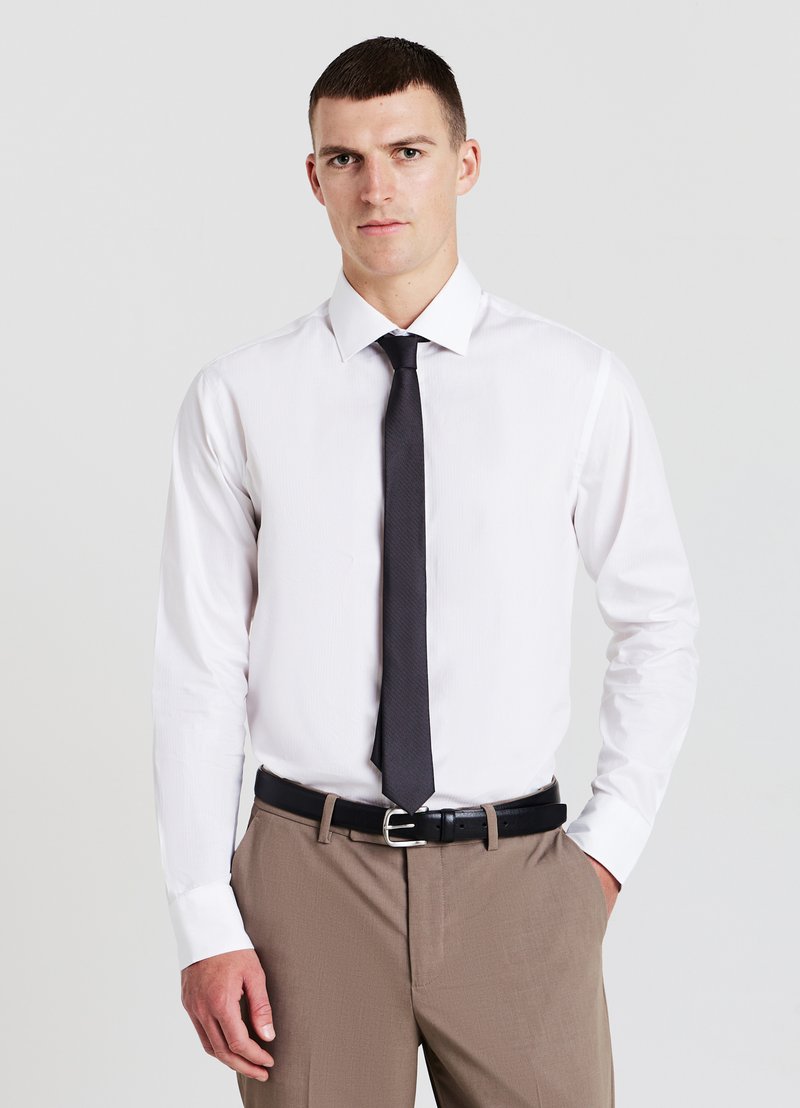 Homme portant une chemise blanche, une cravate noire et un pantalon beige avec une ceinture noire, se tenant devant un fond clair uni.