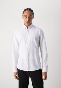 Camisa blanca de manga larga en tejido ligero con cuello abotonado, que presenta una superficie ligeramente texturizada y dobladillo curvado. Combinada con pantalones negros.