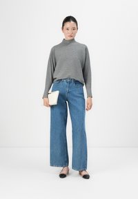 Woman wearing a gray turtleneck sweater, wide-leg blue jeans, two-tone flat shoes, holding a white clutch against a plain white background.