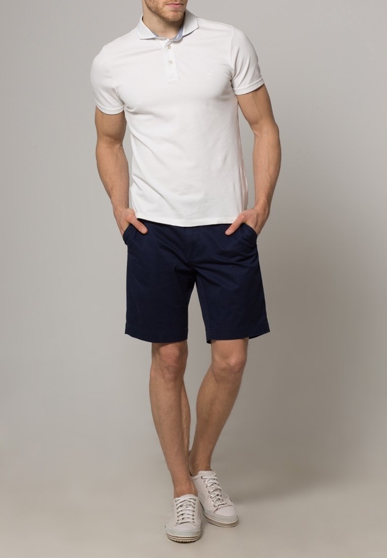 Valkoinen polo-paita, tummansiniset shortsit ja valkoiset lenkkarit. Polossa on klassinen kaulus ja napit. Shortseissa on sivutaskut ja sileä kangas.