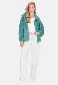 Azurově modrá fleecová bunda s vysokým límcem, dlouhými rukávy a elastickými manžetami, v kombinaci s bílou košilí a světle modrými džíny.