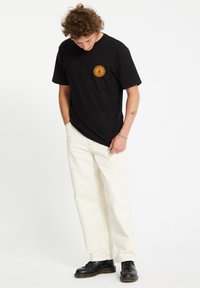 Sort T-shirt med korte ærmer og et rundt orange grafisk motiv på brystet, parret med løs pasform i kremfarvede bukser og sorte sko.
