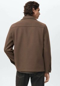 Man met bruin golvend haar, gekleed in een bruine jas en zwarte jeans, die met zijn rug naar de camera staat tegen een effen lichte achtergrond.