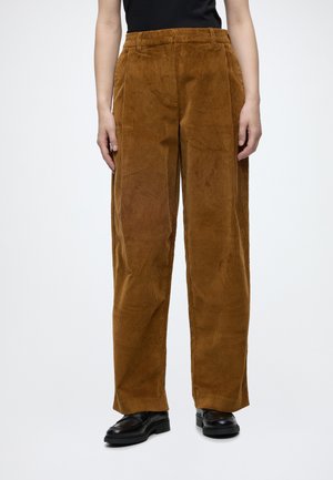 Personne portant un pantalon en velours côtelé marron ample associé à des mocassins noirs et un haut noir sur un fond clair uni.