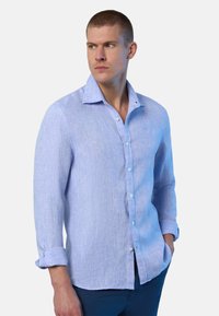 Uomo con capelli corti che indossa una camicia blu chiara a maniche lunghe con i polsini arrotolati e pantaloni scuri, che guarda verso sinistra.