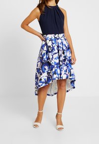 Dopasowany granatowy top bez rękawów z plisowanym dekoltem, zestawiony z długością mini z przodu i maxi z tyłu, w kwiatowy wzór w odcieniach niebieskiego i białego, noszony z białymi sandałami.