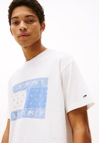 Camiseta de algodón blanca con un gráfico de patrón de bandana azul y blanco con el texto "TOMMY JEANS". Mangas cortas y pequeño logo en la manga.