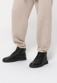 Czarne skórzane buty za kostkę z teksturowanym wykończeniem, okrągłym noskiem i płaską gumową podeszwą, sparowane z luźnymi beżowymi spodniami dresowymi.