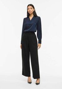 Tumši zila satīna blūze ar v-veida izgriezumu un pogu detaļām, kopā ar augsta vidukļa melnām plātām biksēm un melnām augstpapēžu kurpēm ar zelta akcentiem.