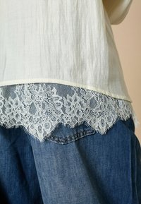 Hvid bluse med en tekstureret overflade, som har en scallop-lace kant i lyseblå. Sæt den sammen med blå denim jeans, der viser lomme detaljer.