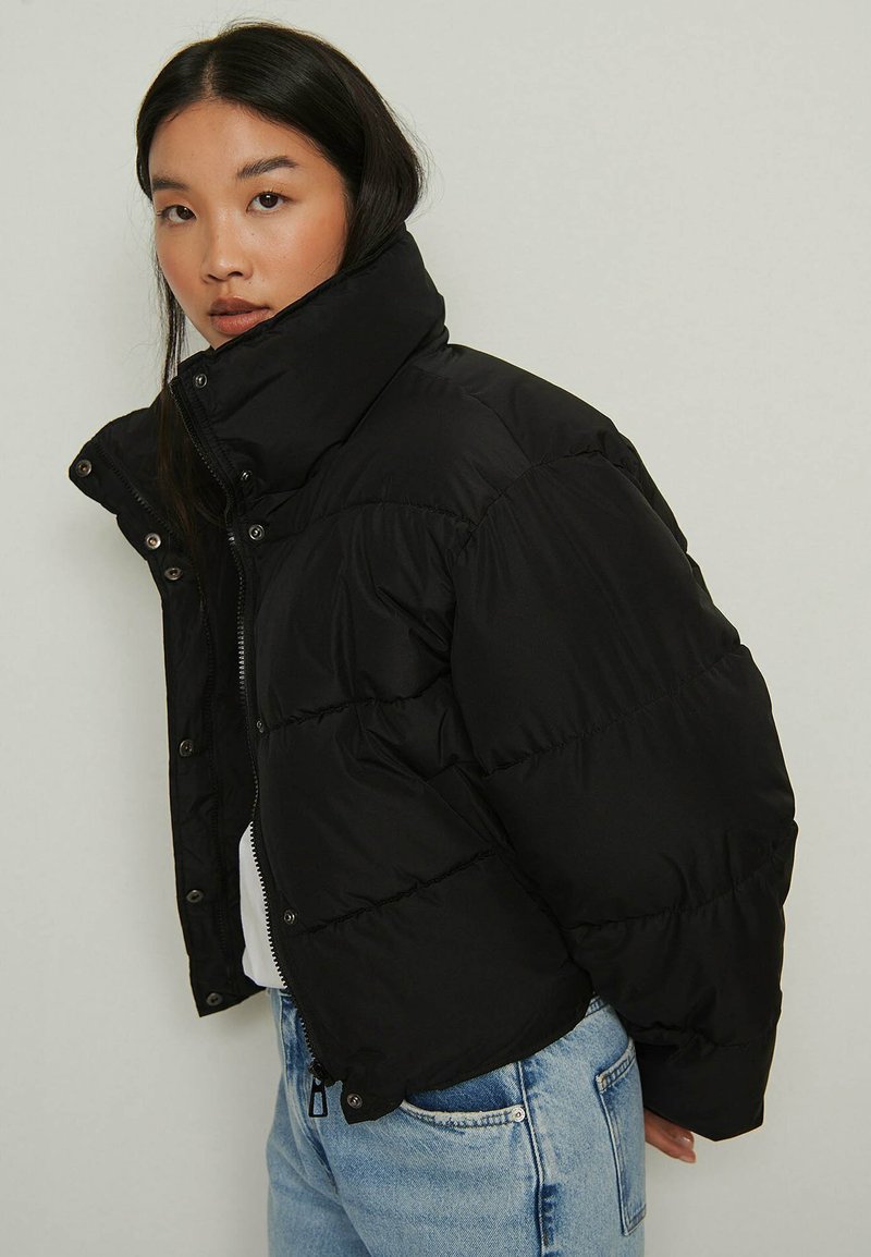 Melna pūkainā jaka ar augstu apkakli, rāvējslēdzēja aizdari un spiedpogu aizdari, ar quilted paneļiem un saīsinātu dizainu.