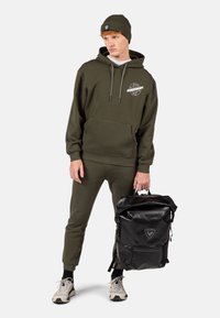 Felpa con cappuccio verde oliva e pantaloni jogger con tasca a marsupio, abbinati a uno zaino nero con logo e spallacci regolabili.