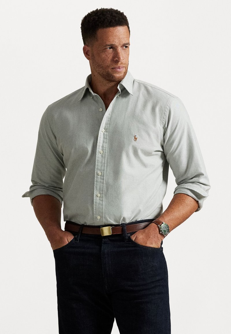 Šviesiai pilkas marškinėlių mygtukais palaidinė su apykakle, puošni mažyte siuvinėta emblema krūtinėje, derinama su tamsiomis džinsinėmis kelnėmis ir rudomis diržais.