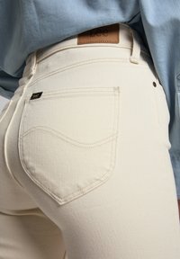 Jeans de denim blanco con tejido texturizado, detalles en el bolsillo trasero y una etiqueta de cuero en la cinturilla que presenta el logo de "Lee".