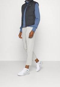 Sort ærmeløs vest over en blå langærmet skjorte, kombineret med lysegrå smalle bukser og hvide sneakers med Nike-logo detaljer.