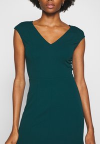 Femme aux cheveux courts et bouclés portant une robe sans manches vert foncé avec un décolleté en V et une taille ajustée sur un fond uni.
