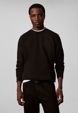 Joven con piel oscura vistiendo una sudadera negra Calvin Klein y pantalones negros sobre un fondo liso gris claro.