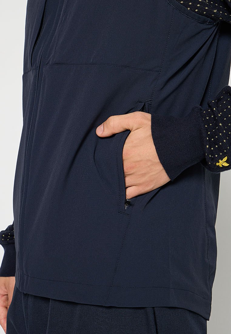 Veste légère bleu marine avec une poche frontale zippée. Équipée de manches noires texturées avec de petits points dorés et des poignets côtelés.
