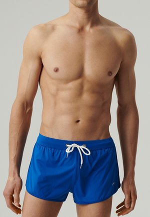 Muskularny mężczyzna w niebieskich szortach kąpielowych z białym ściągaczem, stojący na jednolitym jasnoszarym tle.