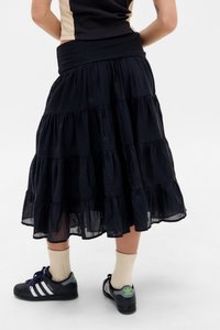 Inimene, kes kannab musta kihilise midi-seeliku, beeže sokke ja musti Adidas'i tossusid valgete triipude ja rohelise logoga, seisab valgel põrandal.