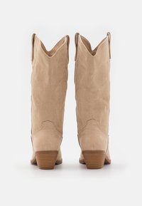 Beige suède cowboylaarzen met decoratieve stiksels, puntige neus en gestapelde hak. Voorzien van trekriemen bovenaan voor eenvoudig aan- en uittrekken.
