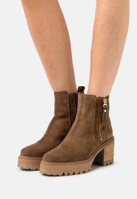 Ruskikankaiset ruskeat nilkkasaappaat, joissa on lohkarekorko, sivuvetoketju ja teksturoitu kumipohja. Korostettu yläosan ympärillä olevalla ompelella.