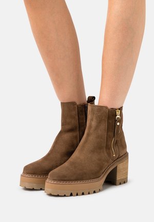 Bottines en daim marron avec un talon bloc, dotées d'une fermeture éclair sur le côté et d'une semelle en caoutchouc texturée. Ornées de coutures autour de la tige.