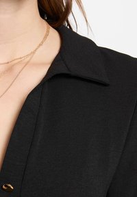Camicia nera con colletto realizzata in tessuto liscio, caratterizzata da un accento di bottone dorato sul lato e collane di metallo dorato sovrapposte contro il colletto.