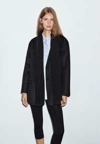 Černý oversized kabát z vlněného materiálu, se zářezovým límcem, předními knoflíky a bočními kapsami, přehozený přes světle modrou košili a černé legíny.