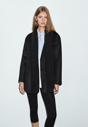 Cappotto oversize nero in misto lana, con revers a lancia, bottoni frontali e tasche laterali, indossato sopra una camicia azzurro chiaro e leggings neri.