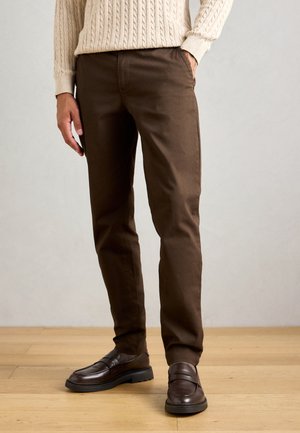 Man wearing brown trousers and dark brown loafers standing on wooden floor with beige cable-knit sweater partially visible.