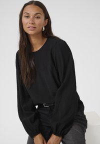 Blouse noire texturée à manches longues, avec un col rond et des poignets élastiques. Portée avec un jean foncé, ornée d'une fine ceinture noire.