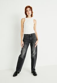Zwarte wijdvallende jeans met een vervaagde afwerking, gecombineerd met een witte geribbelde tanktop. Model staat met handen langs de zij op een effen achtergrond.