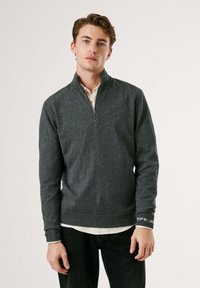 Pelēks zip-up džemperis ar mīkstu tekstūru un garām piedurknēm. Ievērojamais ir apkakles dizains un ribota apakšmala; uz piedurknes ir logo detaļa. Ikdienišķais dizains.