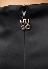 Κοντινή λήψη μαύρου υφάσματος με φερμουάρ που έχει προσαρτημένο ασημένιο γράμμα "HUGO", δείχνοντας λεία υφή και καθαρό σχεδιασμό.
