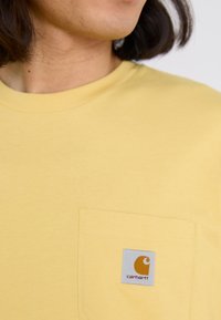 T-shirt en coton jaune avec une poche poitrine et une étiquette logo Carhartt orange sur un fond gris carré. Texture lisse.