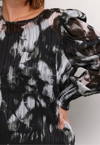 Czarny i biały bluzka z przezroczystego, plisowanego materiału. Posiada bufiaste rękawy oraz abstrakcyjne wzory kwiatowe w różnorodnych odcieniach szarości.