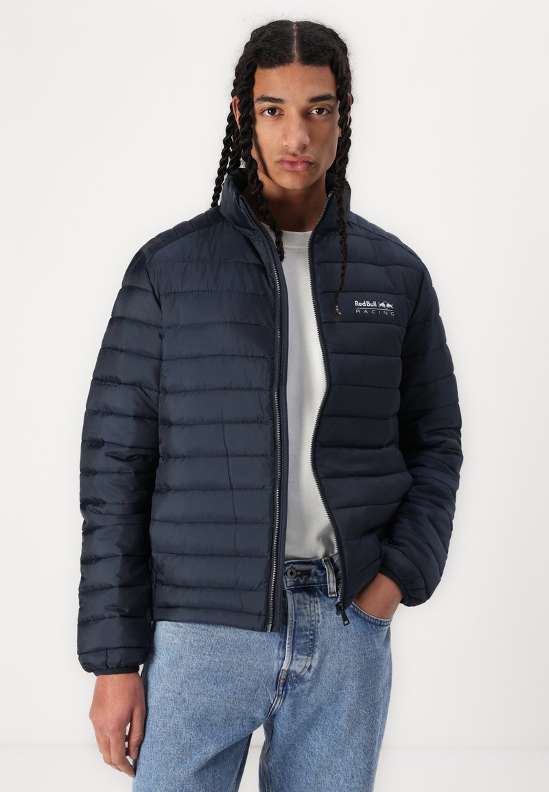 Marine blauwe pufferjack met horizontale stiksels en een voorrits. Bevat het Red Bull Racing-logo op de linkerborst. Textuurmateriaal dat waterafstotend is.