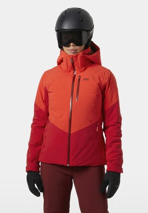 Personne portant une veste de ski rouge, un pantalon bordeaux, des gants noirs, un casque noir et des lunettes de ski noires, debout sur un fond clair.