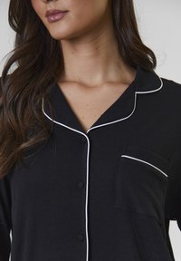 Sort ribbet pyjamas-top med hvide kantdetaljer, der har krave, knaplukning foran og en venstre brystlomme.