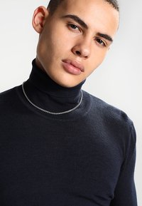Marineblå turtleneck-genser i myk strikket stoff, tettsittende design, med et sølvkjede. Glatt tekstur, minimale synlige sømmer.