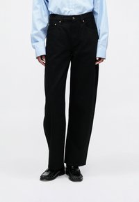 Calças largas pretas de denim, com bolsos e acessórios em tom dourado. Combinadas com uma camisa de botões riscada em azul claro e sapatos pretos.