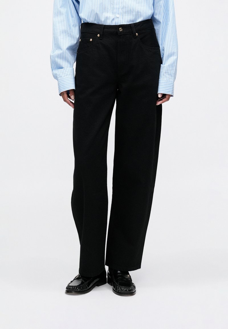 Calças largas pretas de denim, com bolsos e acessórios em tom dourado. Combinadas com uma camisa de botões riscada em azul claro e sapatos pretos.