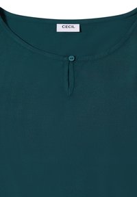 Tealfarbene Bluse mit rundem Halsausschnitt, versehen mit einem kleinen Knopfdetail unterhalb des Halses. Das Etikett trägt die Aufschrift "CECIL" am Kragen. Glatte Stoffoberfläche.